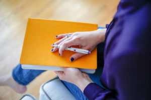 Henkilö istuu oranssi muistikirja ja kynä kädessään.